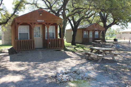Two wooden cabins with porches under trees, a picnic table, and a fire pit in a grassy area.