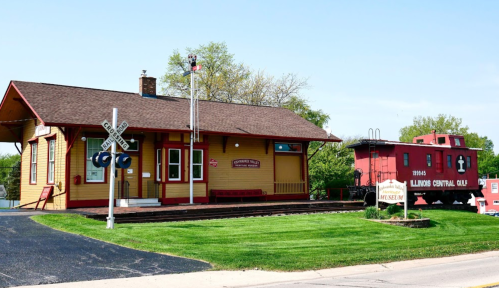 Historic train depot with a red caboose, surrounded by green grass and trees, featuring a railroad crossing sign.