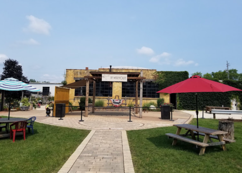 A rustic building with a sign reading "The Warehouse," surrounded by outdoor seating and greenery on a sunny day.