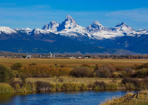 Snow-capped mountains rise above a lush valley, with a river and fields in the foreground under a clear blue sky.