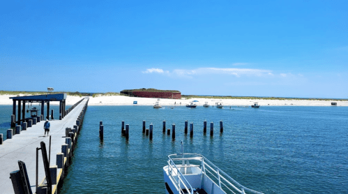 A serene waterfront scene with a pier, boats, and a sandy beach under a clear blue sky.
