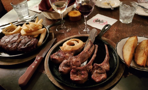 A table setting with a steak, lamb chops, onion rings, fries, and drinks, creating a hearty meal atmosphere.