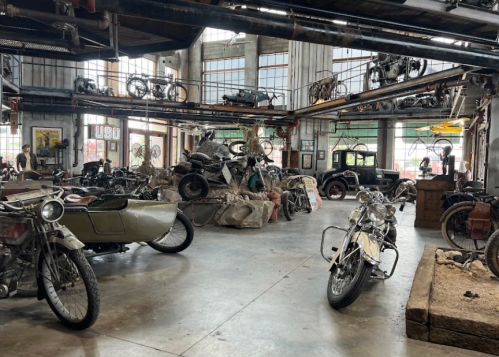 A vintage motorcycle museum filled with various motorcycles, a sidecar, and classic cars in a rustic industrial setting.