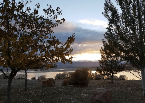 A serene landscape at sunset, featuring trees, a calm lake, and mountains in the background under a cloudy sky.