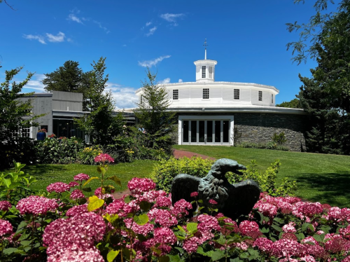 A round white building surrounded by greenery and pink flowers, with a bronze eagle statue in the foreground.
