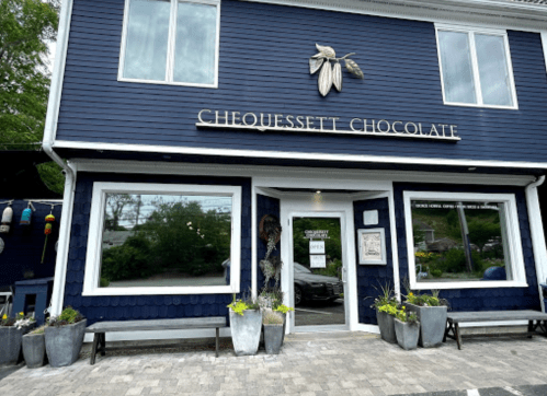 Exterior of Chequessett Chocolate, a blue building with large windows and planters, featuring a sign above the entrance.