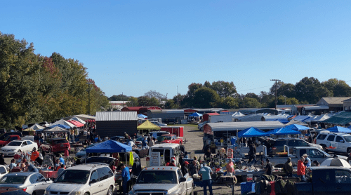 A bustling outdoor market with numerous tents, vendors, and shoppers, surrounded by parked cars under a clear blue sky.