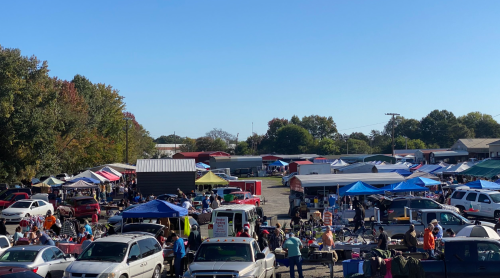 A bustling outdoor market with numerous tents, cars, and people shopping under a clear blue sky.