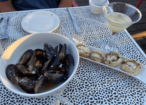 A bowl of mussels in broth, a plate of stuffed mushrooms, and a cocktail on a decorative table.