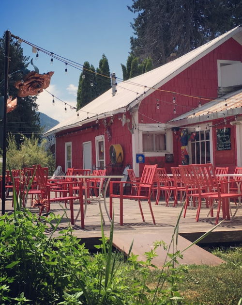 A red building with outdoor seating and string lights, surrounded by greenery and mountains in the background.