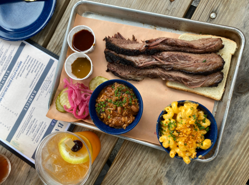 A tray with sliced brisket on bread, sides of macaroni and cheese, baked beans, pickled onions, and sauces.