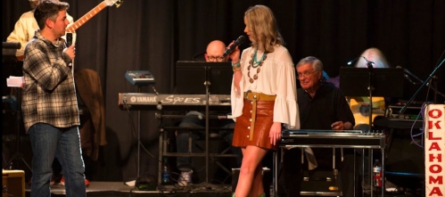 A man and woman perform on stage, with musicians in the background, during a live music event.