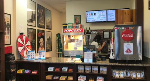 A cozy concession stand with popcorn machine, snacks, and movie posters on the walls. A staff member is serving customers.