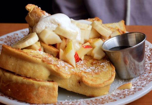 A plate of French toast topped with diced apples, whipped cream, and a small cup of syrup.