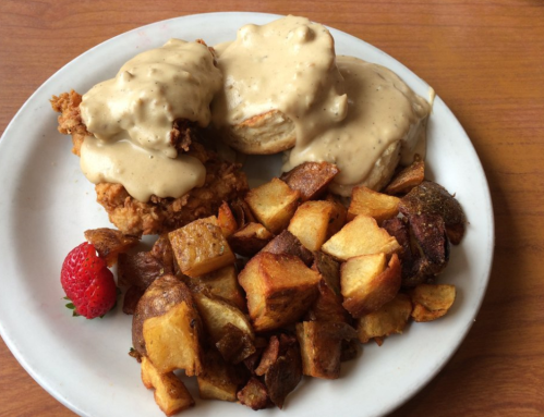 Plate of fried chicken biscuits topped with creamy gravy, served with crispy roasted potatoes and a strawberry.