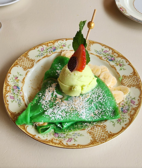 A green crepe topped with ice cream, a strawberry, and mint, served on a decorative plate with banana slices.