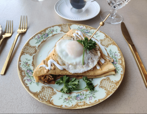 A beautifully plated crepe topped with a fried egg, herbs, and drizzled sauce on an ornate dish with gold utensils.