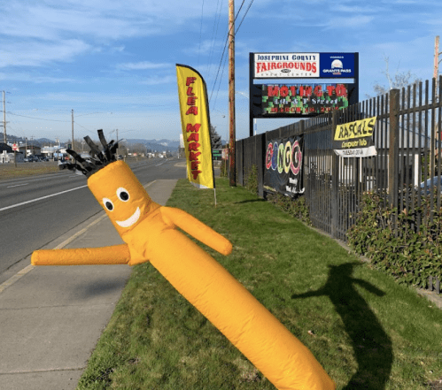 A bright yellow inflatable figure waves outside a flea market near a fairgrounds sign, with a clear blue sky in the background.