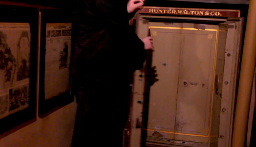 A person in dark clothing stands beside an old safe labeled "HUNTER, WALTON & CO." in a dimly lit room.