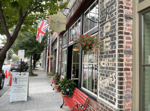 A charming tea room with a British flag, flowers, and a red bench outside a brick building on a city street.