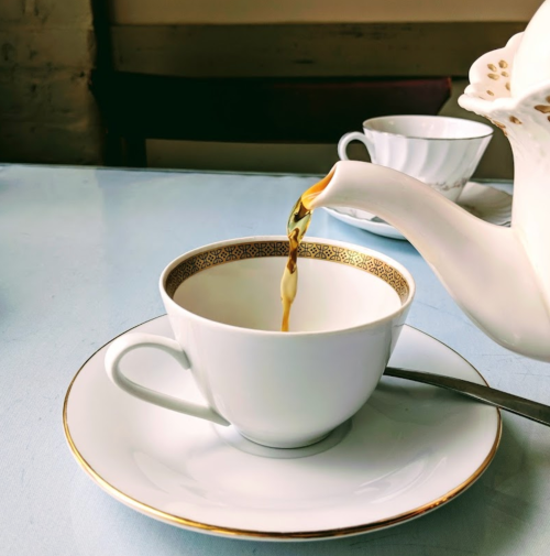 A teapot pours tea into a white cup with a gold rim, set on a light blue table.