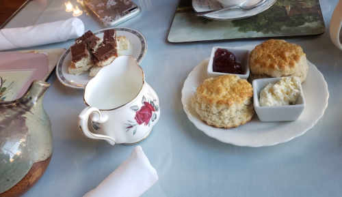 A table setting with scones, cream, jam, a small pitcher, and a plate of dessert squares.