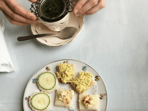 A hand holds a strainer over a cup, while a plate features cucumber slices and small sandwiches with various toppings.