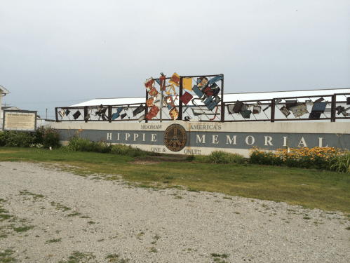 Sign for "Modoc's America's One and Only Hippie Memorial," featuring colorful fabric pieces and a grassy area.
