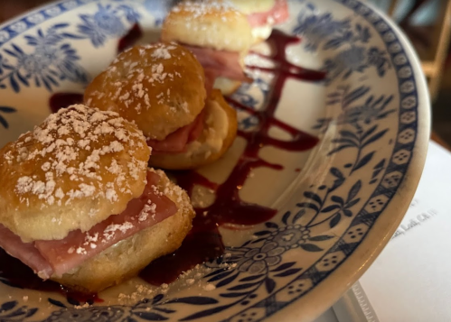 A plate of mini biscuits filled with ham, drizzled with berry sauce and dusted with powdered sugar.