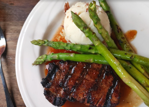 Grilled steak with asparagus and mashed potatoes on a white plate, drizzled with sauce.