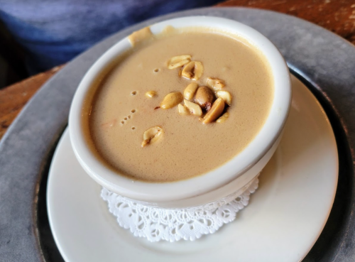 A small bowl of creamy peanut soup topped with peanuts, served on a decorative plate.