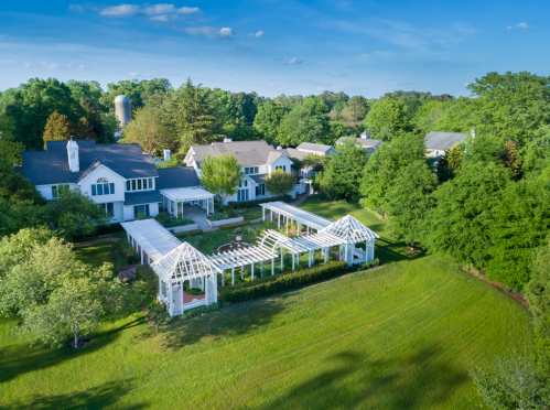 Aerial view of a spacious estate with lush greenery, a white pergola, and several buildings surrounded by trees.