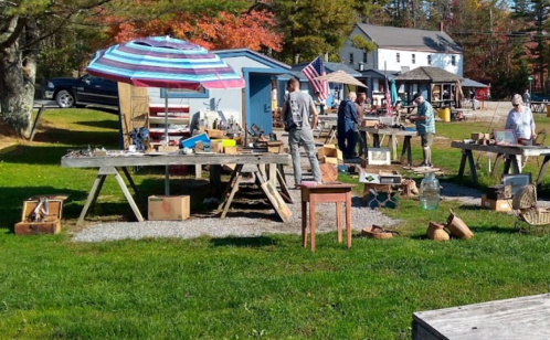 A bustling outdoor market with tables of antiques, people browsing, and colorful umbrellas under a clear sky.