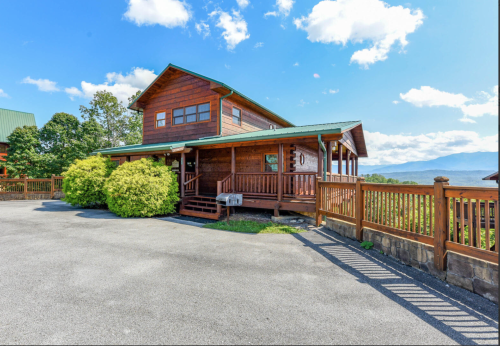 A wooden cabin with a green roof, surrounded by greenery, set against a backdrop of mountains and blue sky.