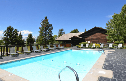 A clear blue swimming pool surrounded by lounge chairs and trees, with a rustic building in the background.