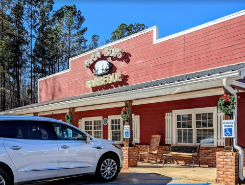Exterior of a barbecue restaurant with a sign, festive decorations, and a parked car in front.