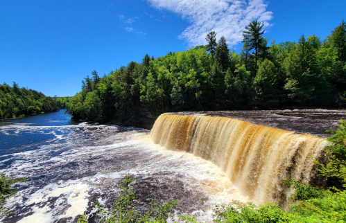 A scenic view of a waterfall cascading into a river, surrounded by lush green trees under a clear blue sky.