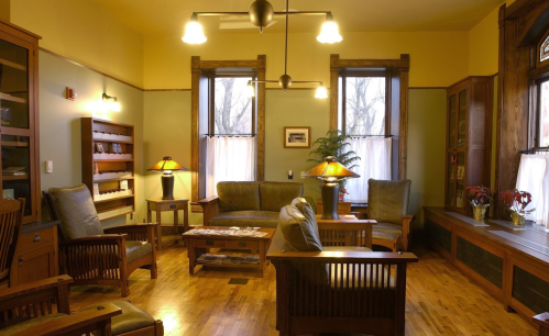 Cozy living room with wooden furniture, large windows, and warm lighting, featuring a plant and magazines on the table.