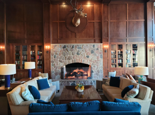 Cozy living room with a stone fireplace, wooden walls, and comfortable seating, featuring a mounted deer head.