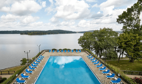A serene outdoor pool with blue water, surrounded by lounge chairs and overlooking a calm lake and trees.