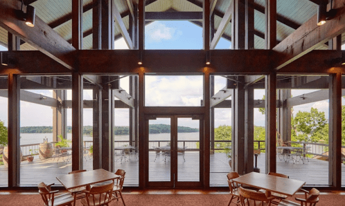 A spacious interior with large windows overlooking a lake, featuring wooden beams and tables set for dining.