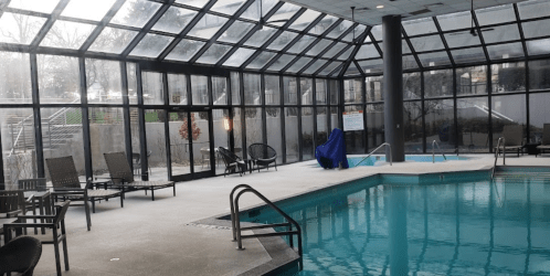 Indoor swimming pool area with glass walls, lounge chairs, and a covered hot tub. Natural light fills the space.