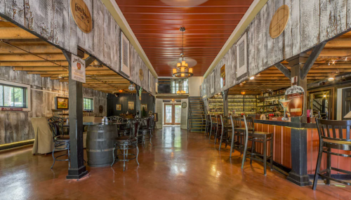 Interior of a rustic bar with wooden beams, a long counter, and seating areas, featuring warm lighting and decor.