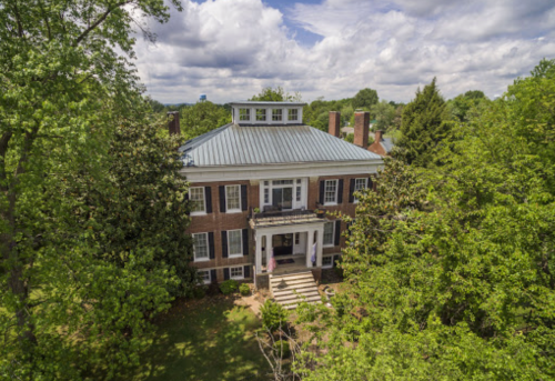 Aerial view of a large brick house with a metal roof, surrounded by lush green trees and a cloudy sky.