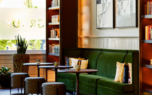 Cozy lounge area with a green sofa, small tables, and bookshelves filled with books, illuminated by natural light.
