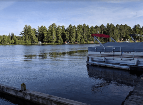 A calm lake surrounded by trees, with a dock and boats moored at the water's edge under a clear sky.