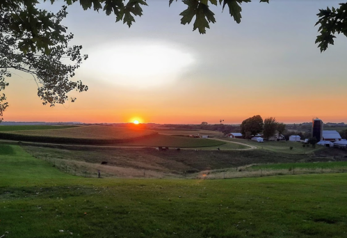 A serene sunset over a rural landscape, with fields, a winding road, and a farm in the distance.