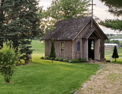 A small wooden chapel surrounded by greenery, with trees and a few animals nearby on a peaceful landscape.