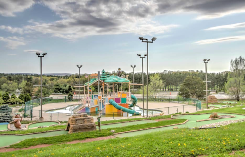Colorful playground with slides and climbing structures, surrounded by green grass and trees under a cloudy sky.