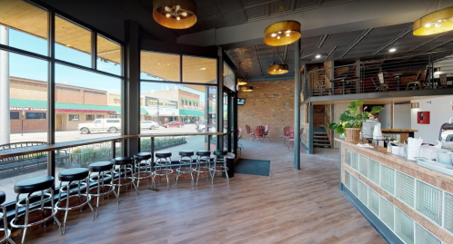 Bright café interior with large windows, wooden floors, and a counter, featuring seating and a view of the street outside.